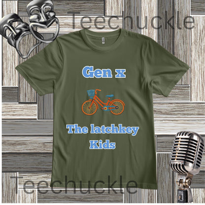 A green graphic t-shirt featuring the text 'Gen x The latchkey Kids' in blue, with an orange bicycle illustration in the center. The t-shirt is displayed on a gray wood-paneled background with vintage microphone and comedy mask props.