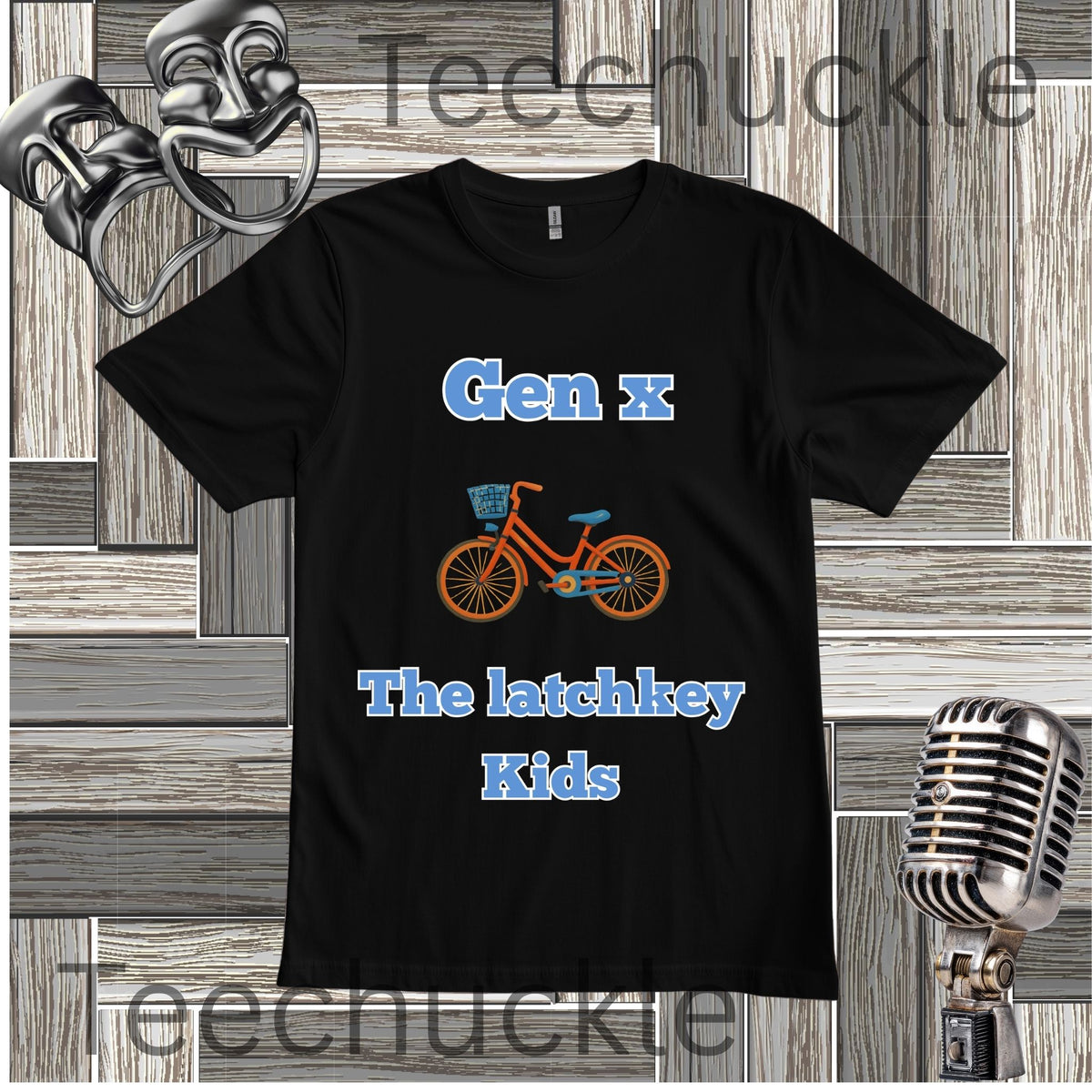 A black short-sleeve graphic t-shirt featuring the text 'Gen x The latchkey Kids' in blue, with an orange and blue bicycle illustration in the center. The t-shirt is displayed on a gray wood-paneled background with decorative props including a silver microphone and theatrical masks