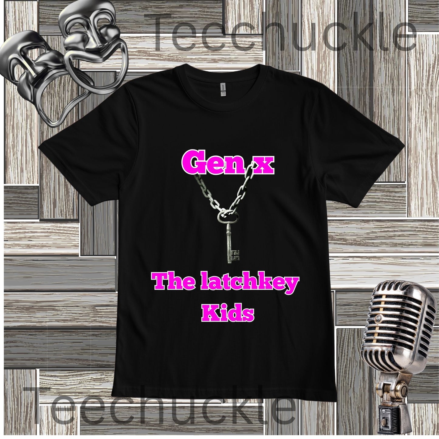 A black short-sleeve graphic T-shirt laid flat on a wood plank background, featuring the text 'Gen X The latchkey Kids' in bright pink and a silver key-and-chain graphic. Decorative theater masks, a vintage microphone, are visible around the shirt.