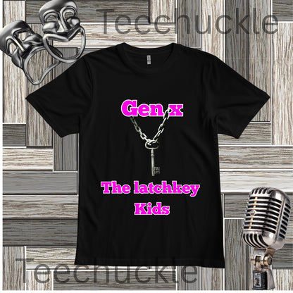 A black short-sleeve graphic T-shirt laid flat on a wood plank background, featuring the text 'Gen X The latchkey Kids' in bright pink and a silver key-and-chain graphic. Decorative theater masks, a vintage microphone, are visible around the shirt.