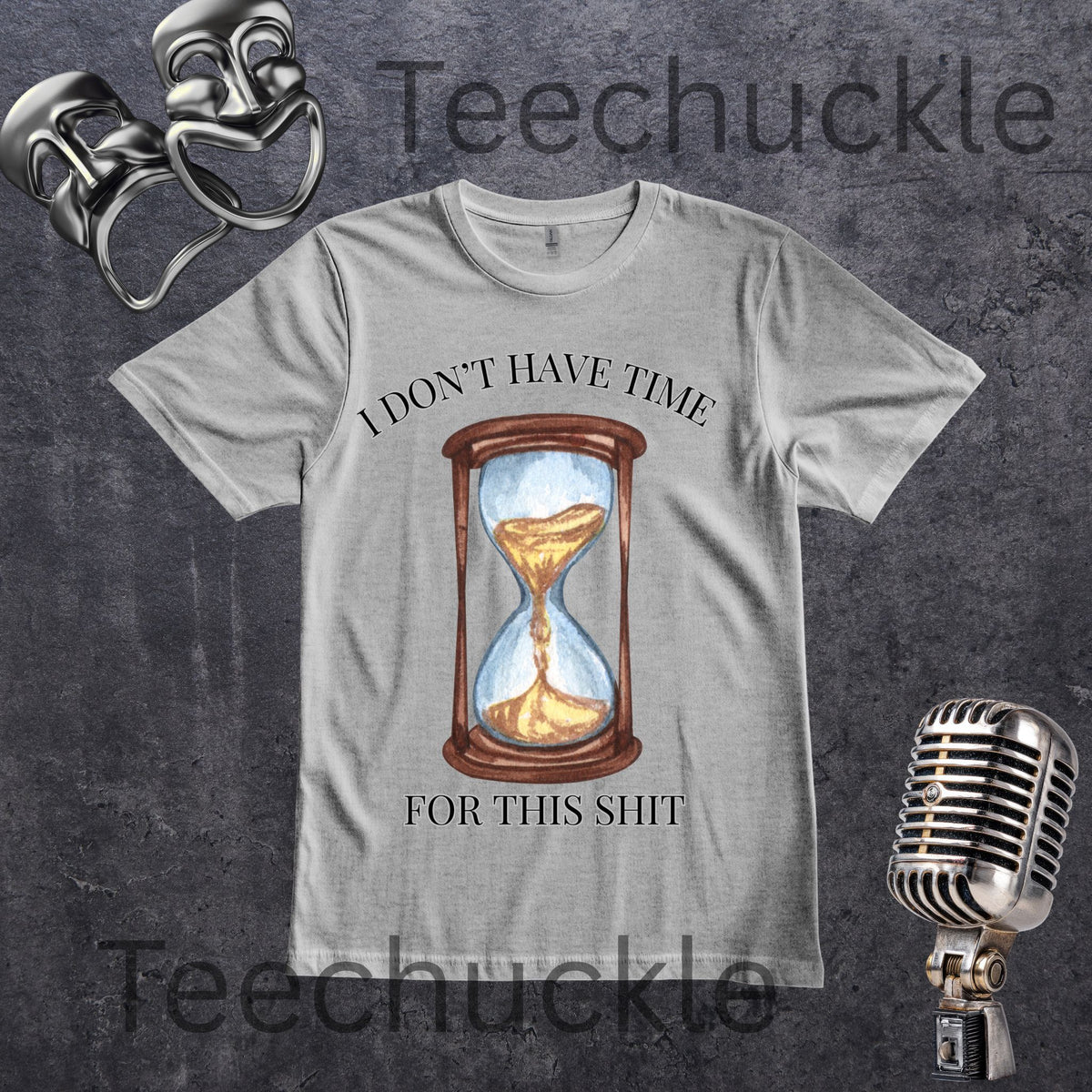 A gray graphic t-shirt featuring a vintage-style printed design of an hourglass with gold sand and the text 'I DON'T HAVE TIME FOR THIS SHIT'. The t-shirt is displayed on a textured dark gray background with vintage props including a microphone, and comedy masks props