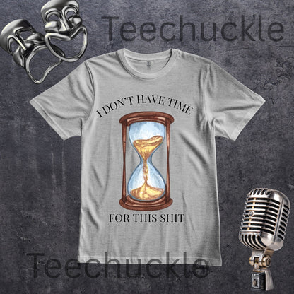 A gray graphic t-shirt featuring a vintage-style printed design of an hourglass with gold sand and the text 'I DON'T HAVE TIME FOR THIS SHIT'. The t-shirt is displayed on a textured dark gray background with vintage props including a microphone, and comedy masks props