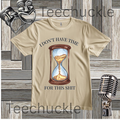 A sand graphic t-shirt featuring a printed illustration of an hourglass with sand flowing through, and the text 'I DON'T HAVE TIME FOR THIS SHIT' in black. The t-shirt is displayed on a gray wood-plank background with vintage microphone props