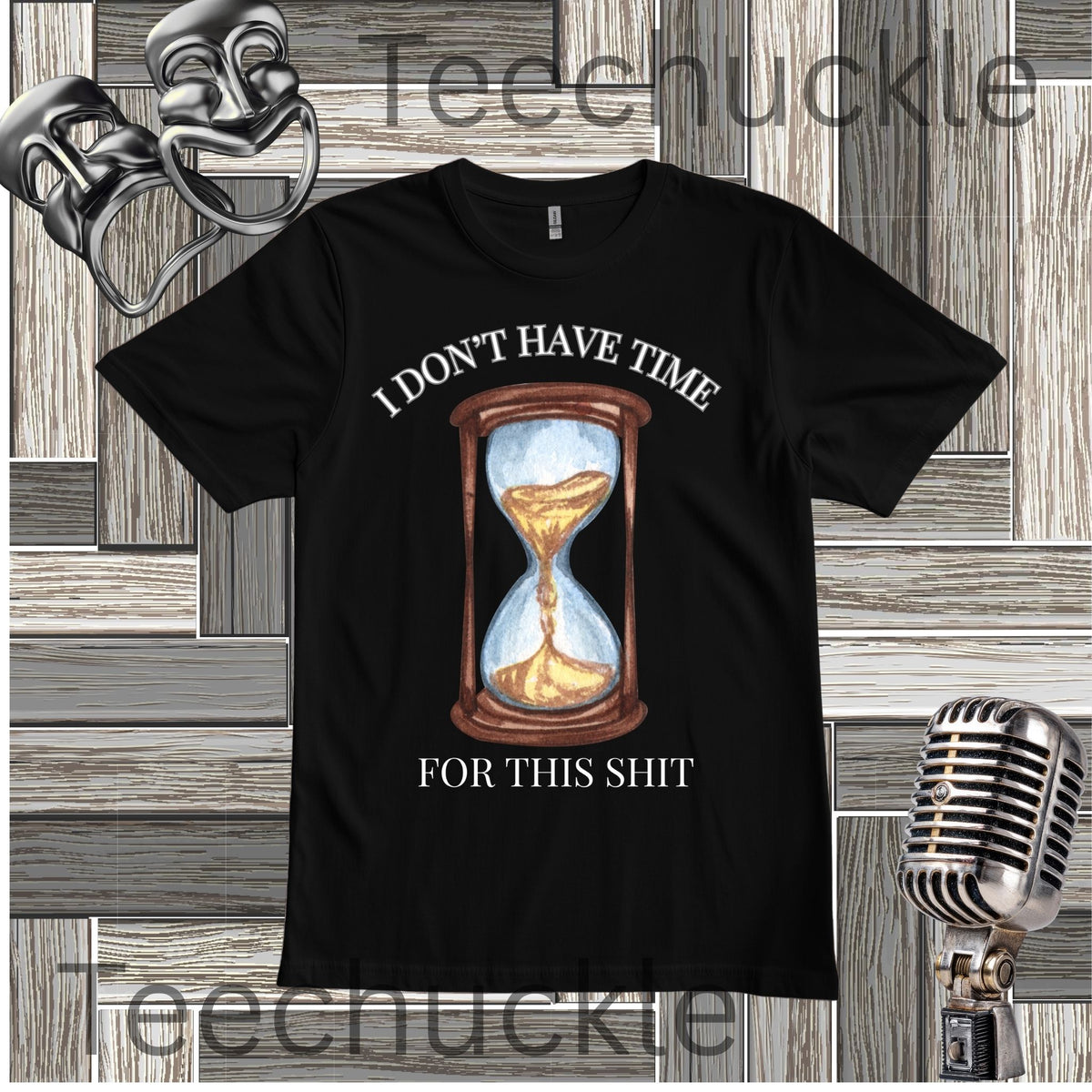 A black graphic t-shirt featuring a vintage-style hourglass illustration with the text 'I DON'T HAVE TIME FOR THIS SHIT' in white. The t-shirt is displayed on a gray wood-paneled background with a silver microphone and comedy masks as props.