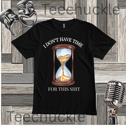 A black graphic t-shirt featuring a vintage-style hourglass illustration with the text 'I DON'T HAVE TIME FOR THIS SHIT' in white. The t-shirt is displayed on a gray wood-paneled background with a silver microphone and comedy masks as props.