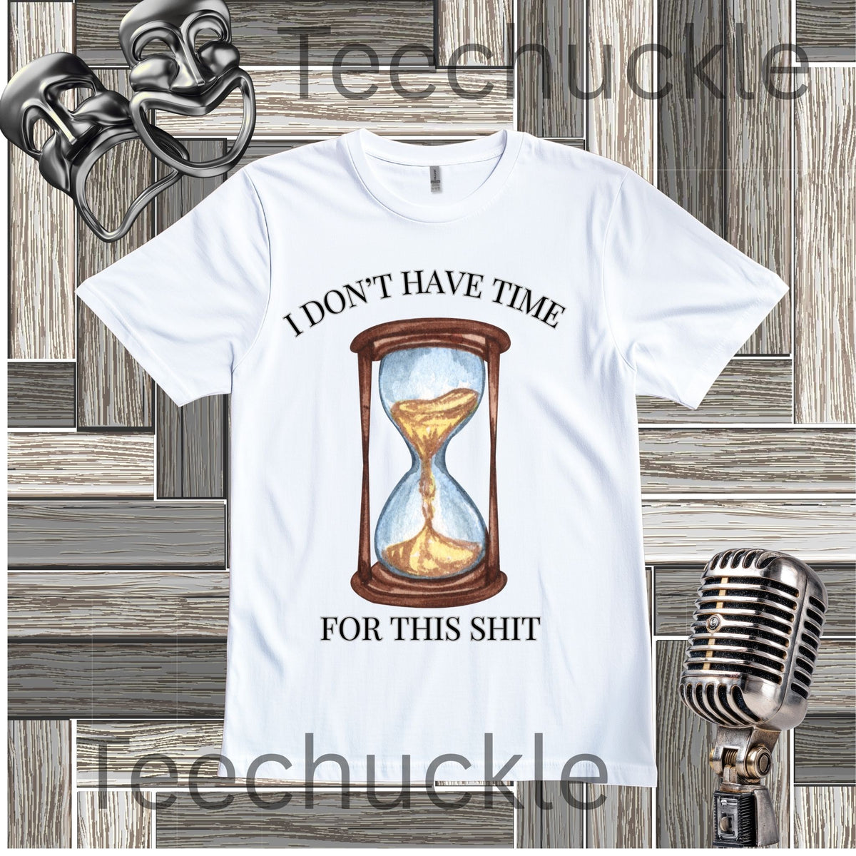 A white graphic t-shirt featuring a watercolor-style hourglass illustration with the text 'I DON'T HAVE TIME FOR THIS SHIT' in black. The shirt is displayed on a gray wood panel background with vintage props, including a silver microphone and two black comedy masks.