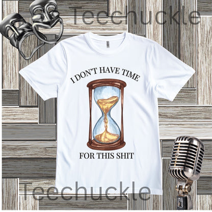 A white graphic t-shirt featuring a watercolor-style hourglass illustration with the text 'I DON'T HAVE TIME FOR THIS SHIT' in black. The shirt is displayed on a gray wood panel background with vintage props, including a silver microphone and two black comedy masks.
