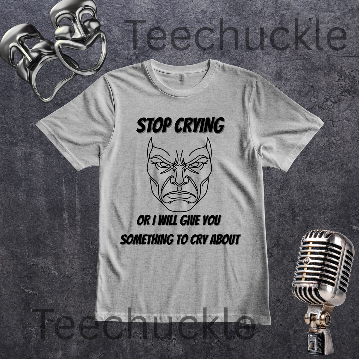 A gray graphic t-shirt featuring a black line art illustration of a angry face with the text 'STOP CRYING OR I WILL GIVE YOU SOMETHING TO CRY ABOUT'. The t-shirt is displayed on a dark textured background with vintage silver boxing gloves, a retro microphone