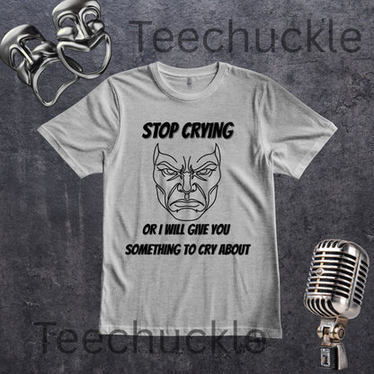 A gray graphic t-shirt featuring a black line art illustration of a angry face with the text 'STOP CRYING OR I WILL GIVE YOU SOMETHING TO CRY ABOUT'. The t-shirt is displayed on a dark textured background with vintage silver boxing gloves, a retro microphone