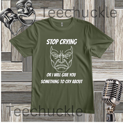 A green graphic t-shirt featuring a white line art illustration of a crying face and the text 'STOP CRYING OR I WILL GIVE YOU SOMETHING TO CRY ABOUT'. The t-shirt is displayed on a gray wood-paneled background with a silver vintage microphone and a silver metal comical mask on either side