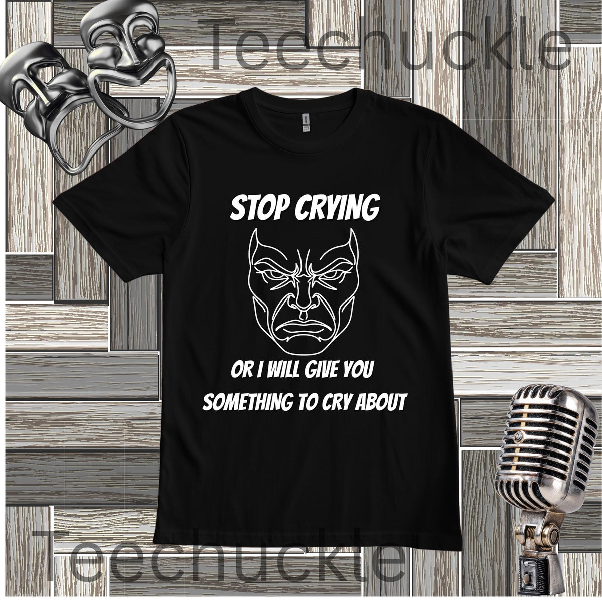 A black short-sleeve graphic T-shirt laid flat on a wood plank background, with white text reading 'STOP CRYING OR I WILL GIVE YOU SOMETHING TO CRY ABOUT' and a white illustrated angry face. Decorative theater-themed props, including a mask, microphone, and backdrop, surround the shirt.