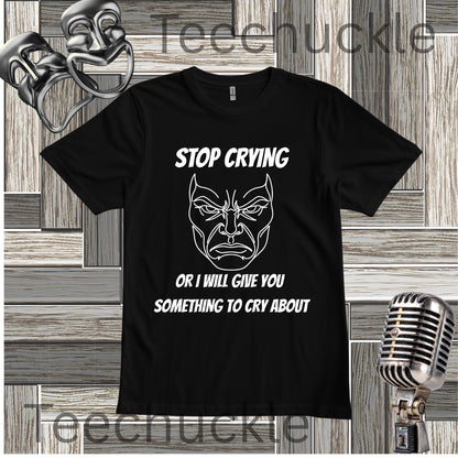 A black short-sleeve graphic T-shirt laid flat on a wood plank background, with white text reading 'STOP CRYING OR I WILL GIVE YOU SOMETHING TO CRY ABOUT' and a white illustrated angry face. Decorative theater-themed props, including a mask, microphone, and backdrop, surround the shirt.