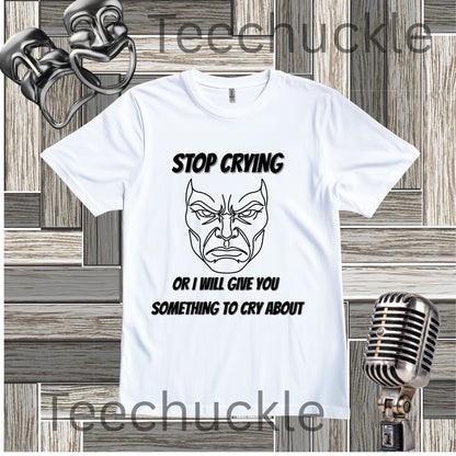 A white graphic t-shirt featuring a black line art illustration of a angry face and the text 'STOP CRYING OR I WILL GIVE YOU SOMETHING TO CRY ABOUT'. The t-shirt is displayed on a gray wood panel background with vintage props, including a silver microphone and two theatrical masks.