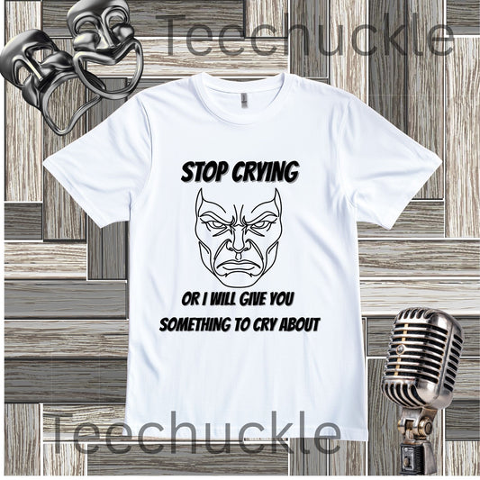 A white graphic t-shirt featuring a black line art illustration of a angry face and the text 'STOP CRYING OR I WILL GIVE YOU SOMETHING TO CRY ABOUT'. The t-shirt is displayed on a gray wood panel background with vintage props, including a silver microphone and two theatrical masks.