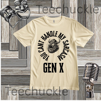 A cream short-sleeve graphic t-shirt with a black circular print featuring a roaring viking man and the text 'YOU CAN'T HANDLE MY SARCASM GEN X'. The shirt is displayed on a rustic wood panel background with vintage-style props, including a silver microphone and a comedy mask