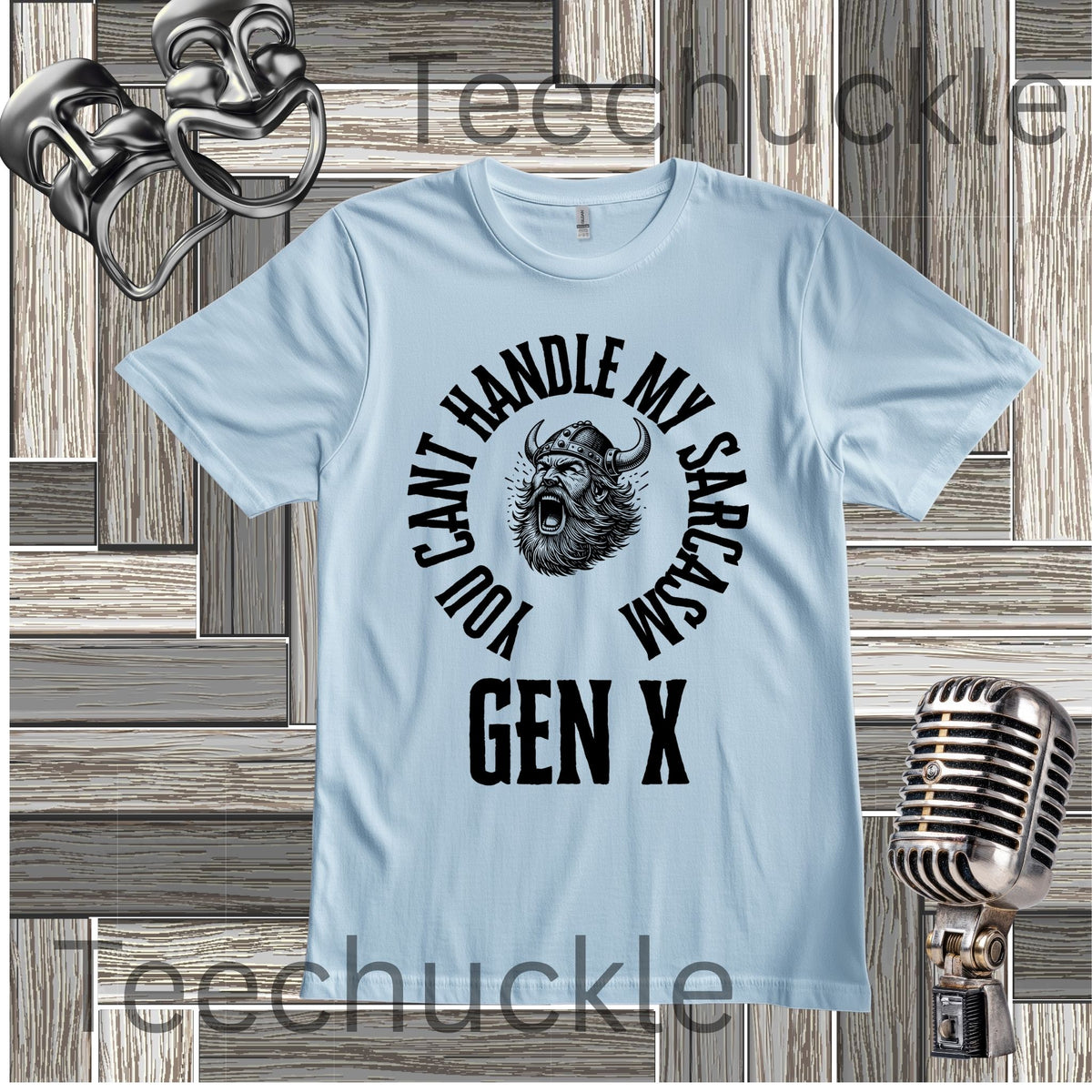 A baby blue short-sleeve graphic t-shirt with a black circular print featuring a roaring viking man and the text 'YOU CAN'T HANDLE MY SARCASM GEN X'. The shirt is displayed on a rustic wood panel background with vintage-style props, including a silver microphone and a comedy mask