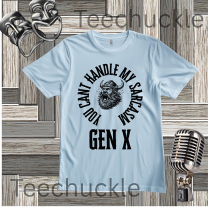 A baby blue short-sleeve graphic t-shirt with a black circular print featuring a roaring viking man and the text 'YOU CAN'T HANDLE MY SARCASM GEN X'. The shirt is displayed on a rustic wood panel background with vintage-style props, including a silver microphone and a comedy mask