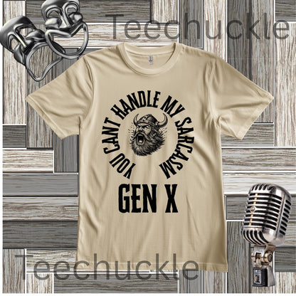 A sand graphic t-shirt featuring a black print of a horned viking head with the text 'YOU CAN'T HANDLE MY SARCASM GEN X' arched around it. The t-shirt is displayed on a gray wood plank background with vintage microphone and headset props
