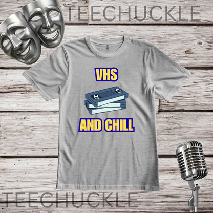 A gray crewneck t-shirt with a graphic of stacked VHS tapes and the text 'VHS AND CHILL' in yellow with blue outlines, displayed on a rustic wood background with a vintage microphone and comedy masks.