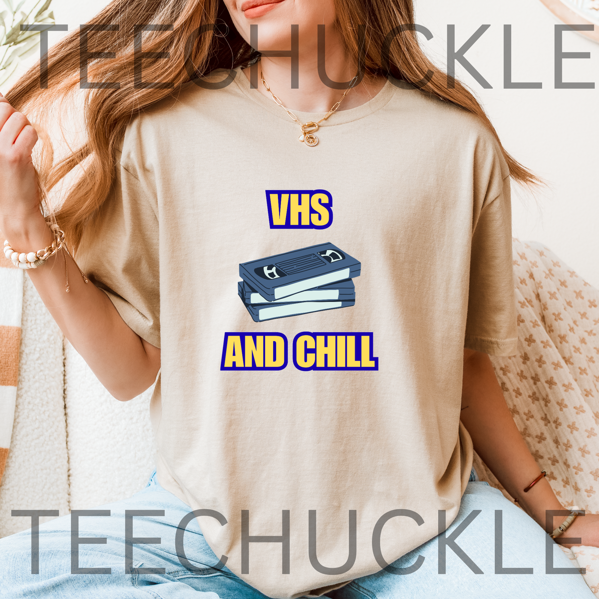 A woman wearing a sand graphic t-shirt with a blue and yellow 'VHS AND CHILL' design, sitting in a cozy indoor setting. 