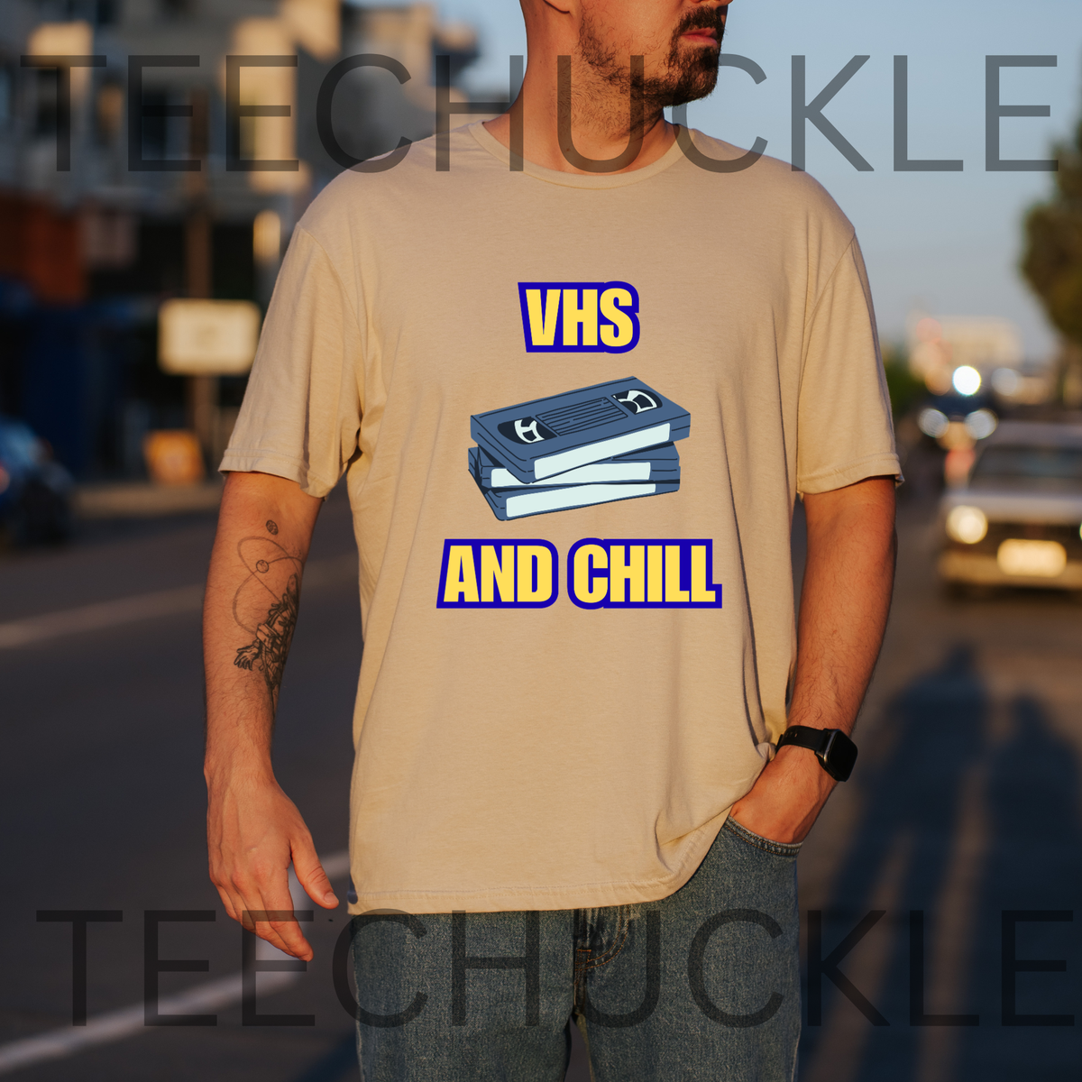 A man wearing a sand graphic t-shirt with a 'VHS AND CHILL' design featuring a blue VHS tape illustration, standing outdoors on a street at sunset. 