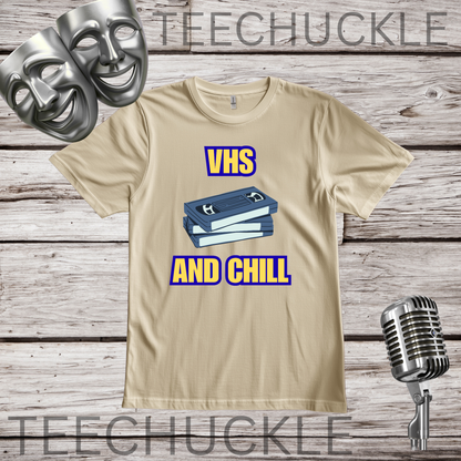 A sand graphic t-shirt featuring a blue VHS tape illustration with the text 'VHS AND CHILL' in yellow and blue. The t-shirt is displayed on a rustic wood background with silver comedy and tragedy masks, a vintage microphone.