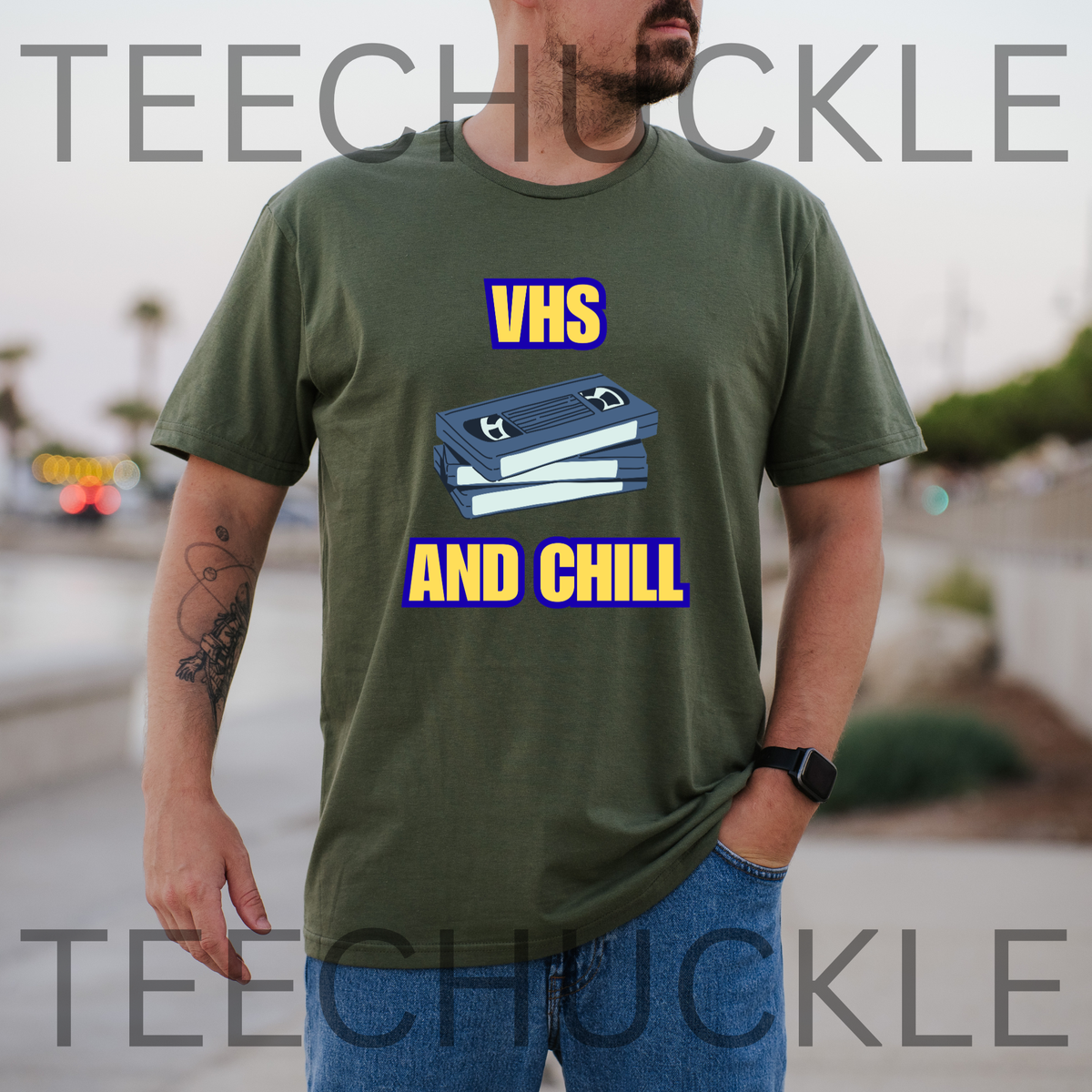 A man wearing a green graphic t-shirt with a blue VHS tape illustration and the text 'VHS AND CHILL' in yellow and blue. The background is a blurred outdoor street scene