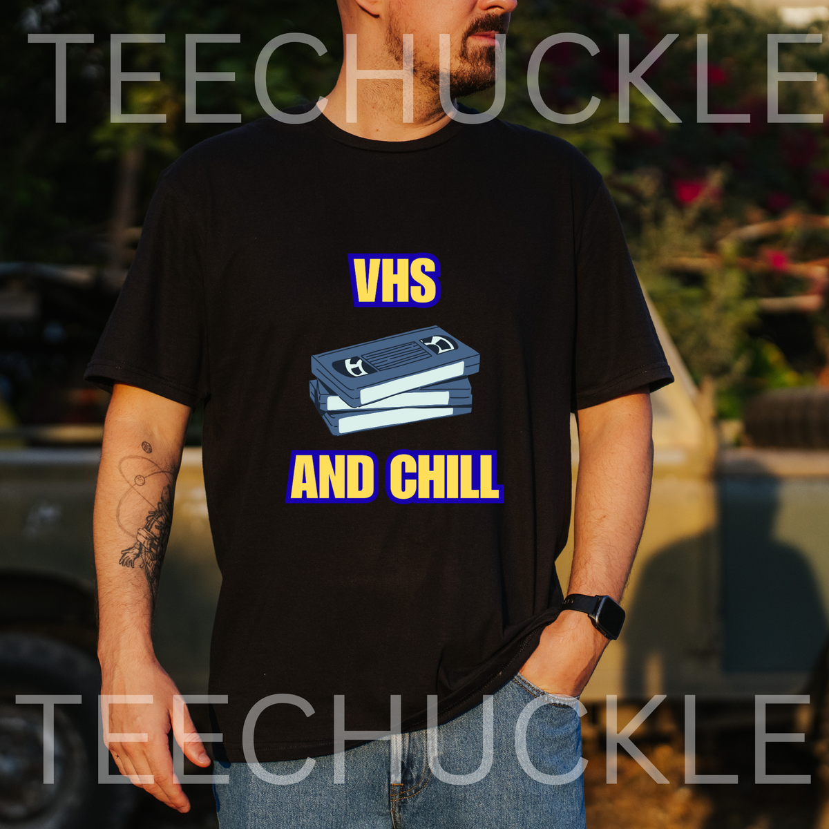 A man wearing a black graphic t-shirt with a 'VHS AND CHILL' design featuring a blue cassette tape illustration, set against a blurred outdoor background