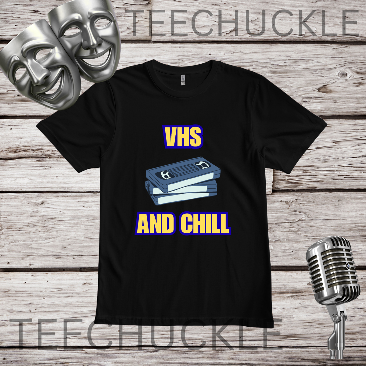A black short-sleeve graphic T-shirt laid flat on a rustic wood plank background, with two silver theater masks, a vintage microphone. The shirt graphic shows stacked VHS tapes with the words “VHS AND CHILL” in yellow with a blue outline.