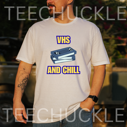 A man wearing a white graphic t-shirt with a blue VHS tape illustration and the text 'VHS AND CHILL' in yellow and blue. The background is a blurred outdoor setting 