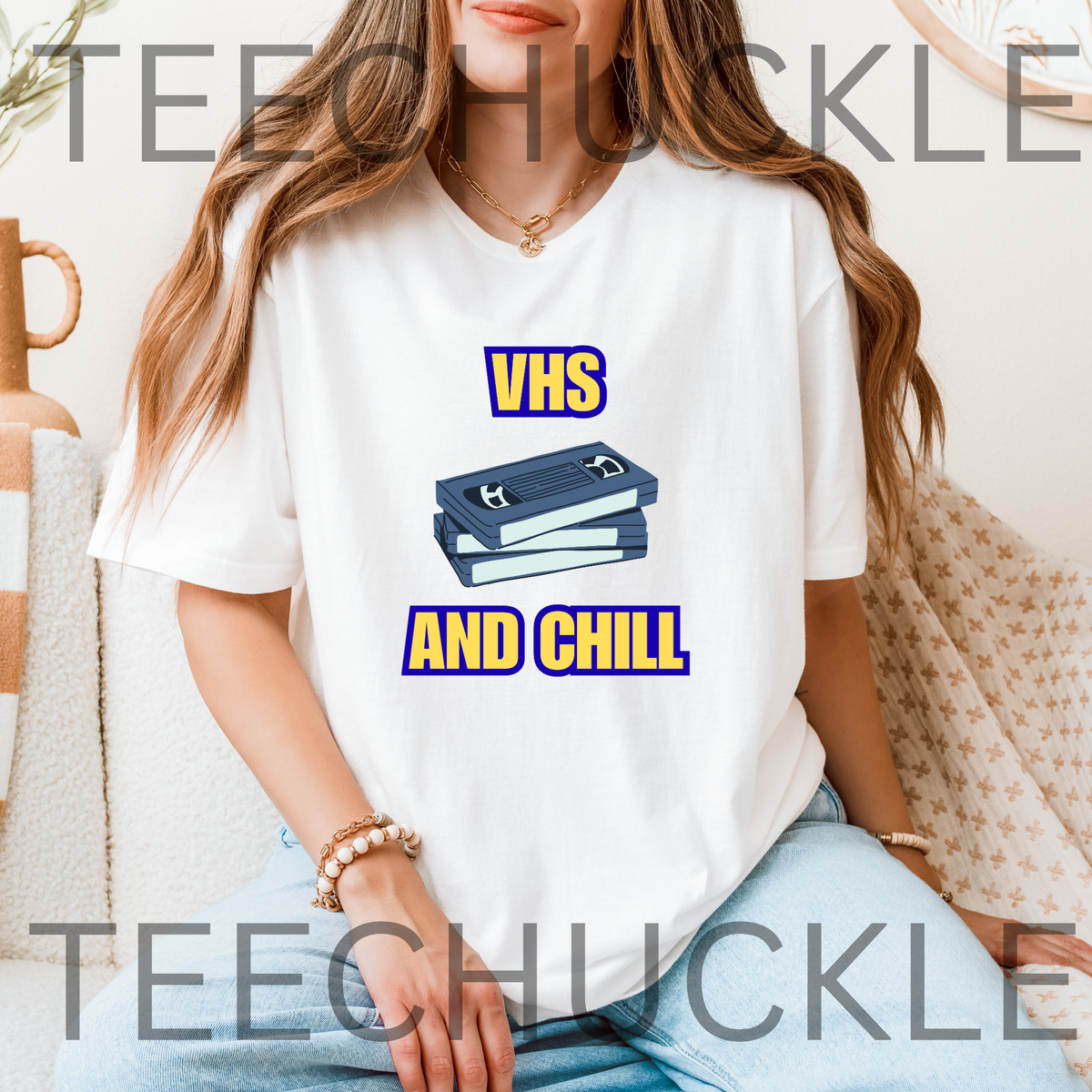 A woman wearing a white graphic t-shirt with a blue and yellow 'VHS AND CHILL' design, sitting in a cozy indoor setting. 