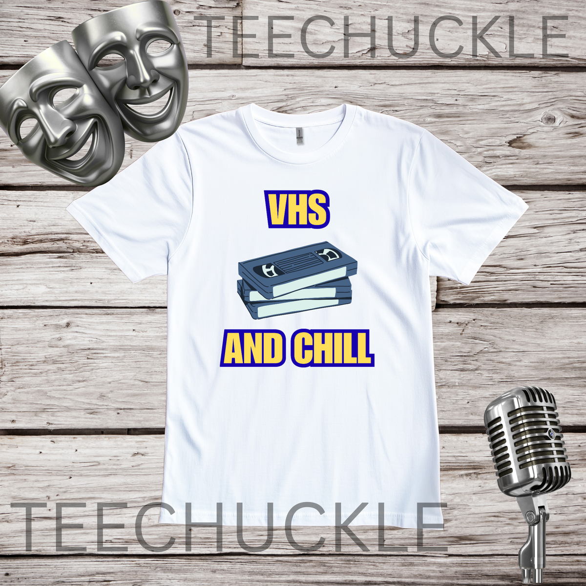 A white graphic t-shirt displayed on a rustic wood background, featuring a 'VHS AND CHILL' design with two VHS tapes. There is two silver theatrical masks and a vintage microphone prop