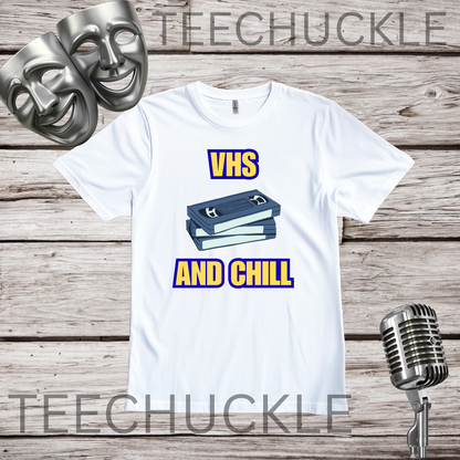 A white graphic t-shirt displayed on a rustic wood background, featuring a 'VHS AND CHILL' design with two VHS tapes. There is two silver theatrical masks and a vintage microphone prop