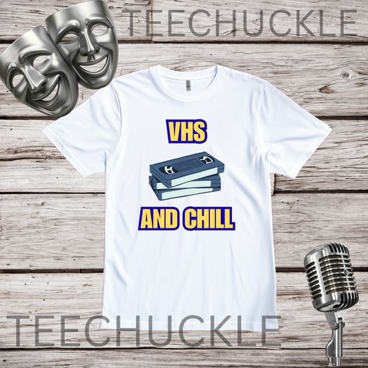 A white graphic t-shirt displayed on a rustic wood background, featuring a 'VHS AND CHILL' design with two VHS tapes. There is two silver theatrical masks and a vintage microphone prop
