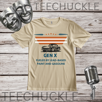 A sand graphic t-shirt featuring a retro design with a black car, orange and blue stripes, and the text 'GEN X FUELED BY LEAD-BASED PAINT AND GASOLINE'. The shirt is displayed on a rustic wood background with silver theater masks and a vintage microphone.