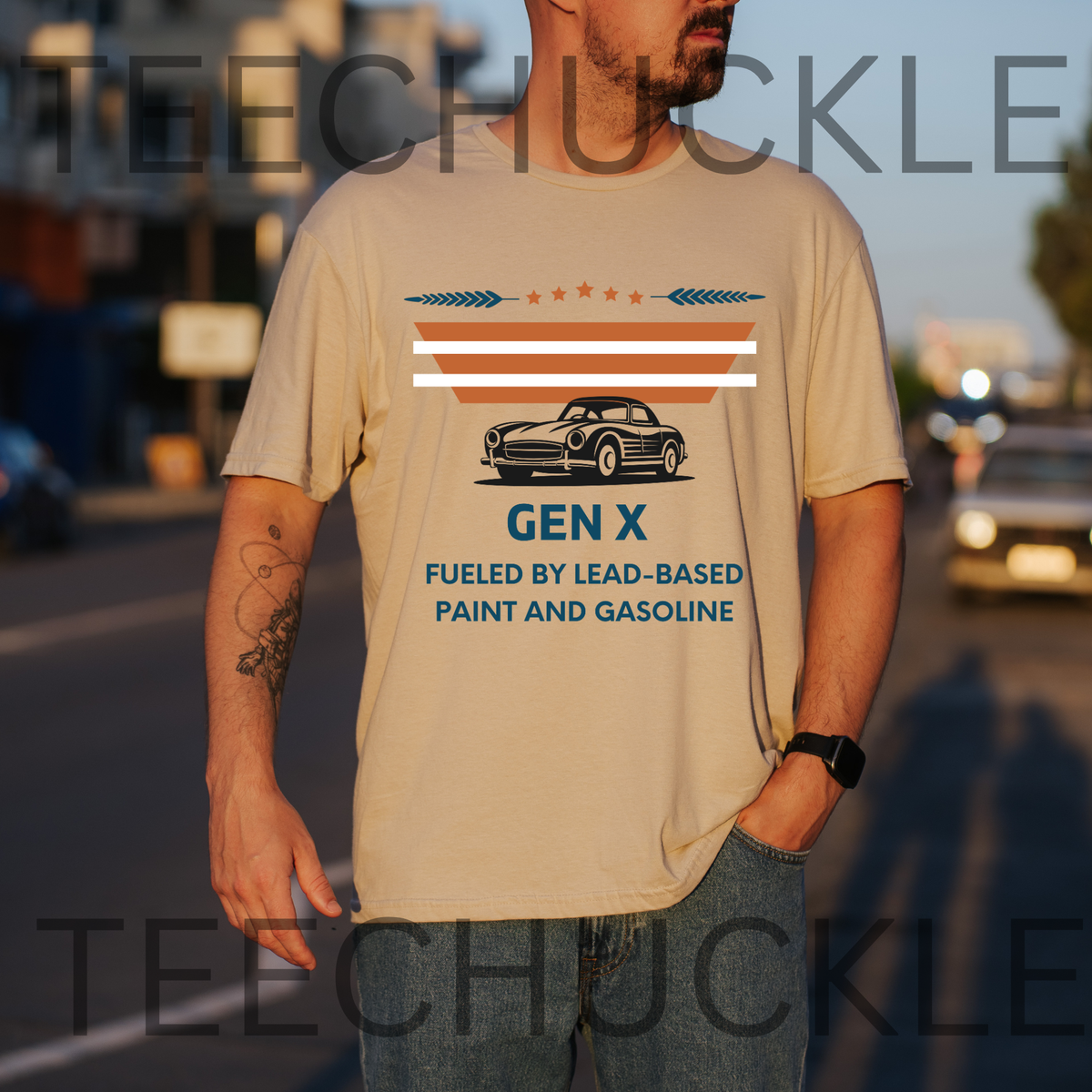 A man wearing a sand graphic t-shirt with a vintage Gen X car design and text, standing on a blurred street at sunset. The shirt features a retro striped design and a black car illustration with the text 'GEN X FUELED BY LEAD-BASED PAINT AND GASOLINE'.