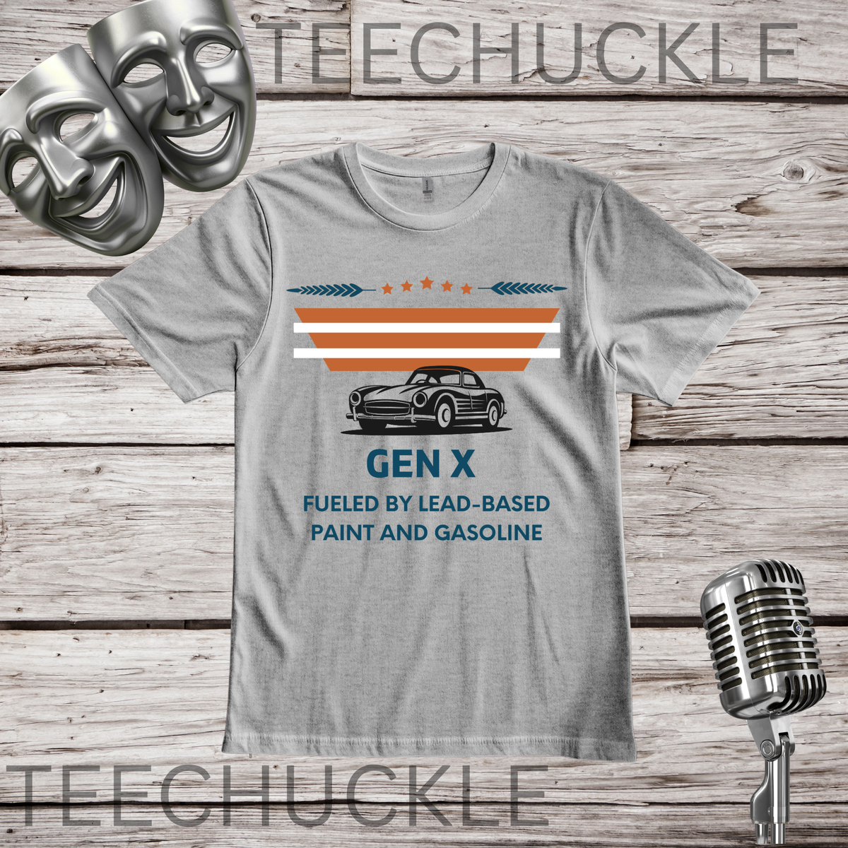 A gray graphic t-shirt displayed flat on a rustic wood background, featuring a vintage car illustration and the text 'GEN X FUELED BY LEAD-BASED PAINT AND GASOLINE'. The shirt is styled with a silver microphone and comedy masks, 