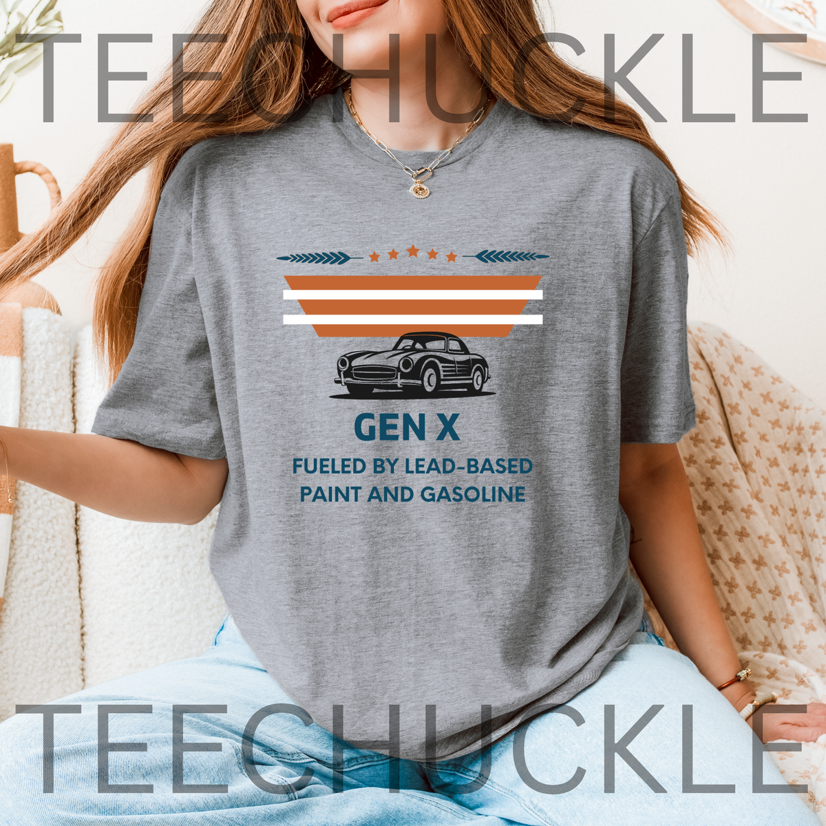 A woman wearing a gray graphic t-shirt with a retro Gen X car design and text, sitting in a cozy indoor setting. The t-shirt features a black vintage car graphic with orange and white stripes above it, and the text 'GEN X FUELED BY LEAD-BASED PAINT AND GASOLINE' printed below.
