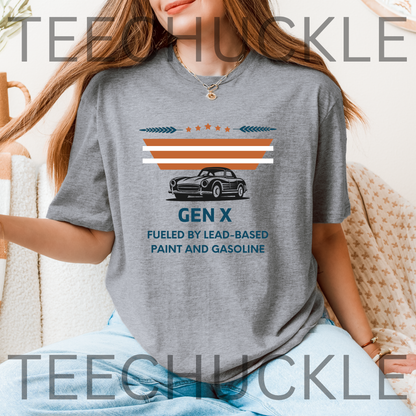 A woman wearing a gray graphic t-shirt with a retro Gen X car design and text, sitting in a cozy indoor setting. The t-shirt features a black vintage car graphic with orange and white stripes above it, and the text 'GEN X FUELED BY LEAD-BASED PAINT AND GASOLINE' printed below.