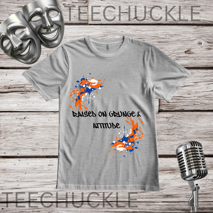 A gray crewneck t-shirt with a graphic design featuring orange and blue paint splatters and the text 'RAISED ON GRUNGE & ATTITUDE' in black. The shirt is displayed on a rustic wood background with decorative props including a silver microphone, comedy masks