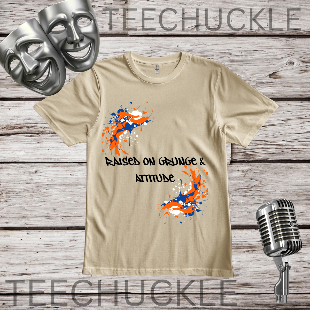 A sand graphic t-shirt displayed on a rustic wood background, featuring orange and blue paint splatters and the text 'RAISED ON GRUNGE & ATTITUDE'. The shirt is decorated with silver comedy masks on the top left, a vintage microphone on the bottom right