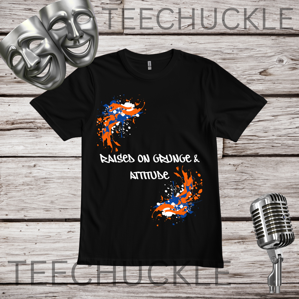 A black short-sleeve graphic T-shirt laid flat on a rustic wood background, with theatrical masks, a vintage microphone. The shirt has a white slogan reading 'Raised on Grunge & Attitude' between two orange, white, and blue paint-splatter graphics.