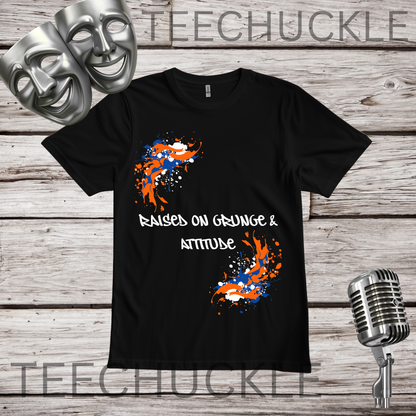 A black short-sleeve graphic T-shirt laid flat on a rustic wood background, with theatrical masks, a vintage microphone. The shirt has a white slogan reading 'Raised on Grunge & Attitude' between two orange, white, and blue paint-splatter graphics.