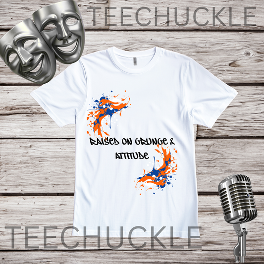 A white graphic t-shirt displayed on a rustic wood background, featuring orange and blue paint splatters and the text 'RAISED ON GRUNGE & ATTITUDE'. The shirt is styled with silver comedy and tragedy masks and a vintage microphone.