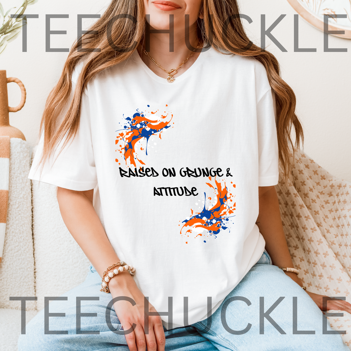 A woman wearing a white graphic T-shirt with orange and blue paint splatters and the text 'RAISED ON GRUNGE & ATTITUDE'. The photo is taken in a cozy indoor setting with a couch and blanket in the background.
