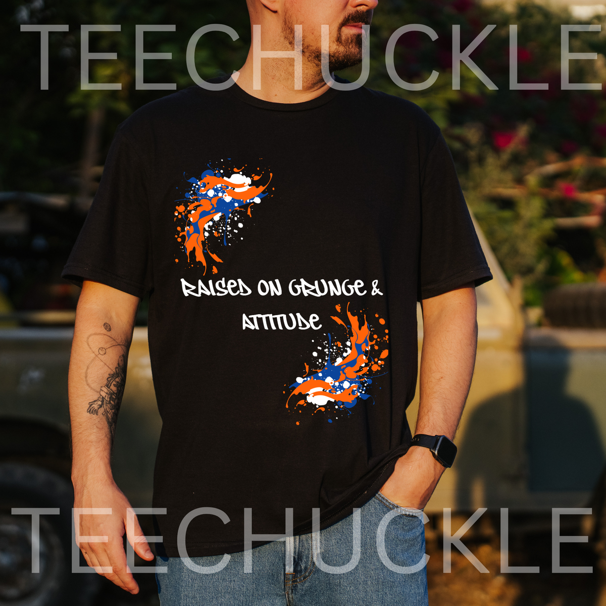 A man wearing a black graphic t-shirt with orange, white, and blue paint splatters and the text 'RAISED ON GRUNGE & ATTITUDE'. The background is an outdoor setting with a blurred truck and foliage
