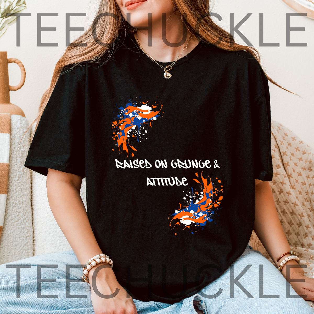 A woman wearing a black graphic t-shirt with orange, blue, and white paint splatters and the text 'RAISED ON GRUNGE & ATTITUDE'. 