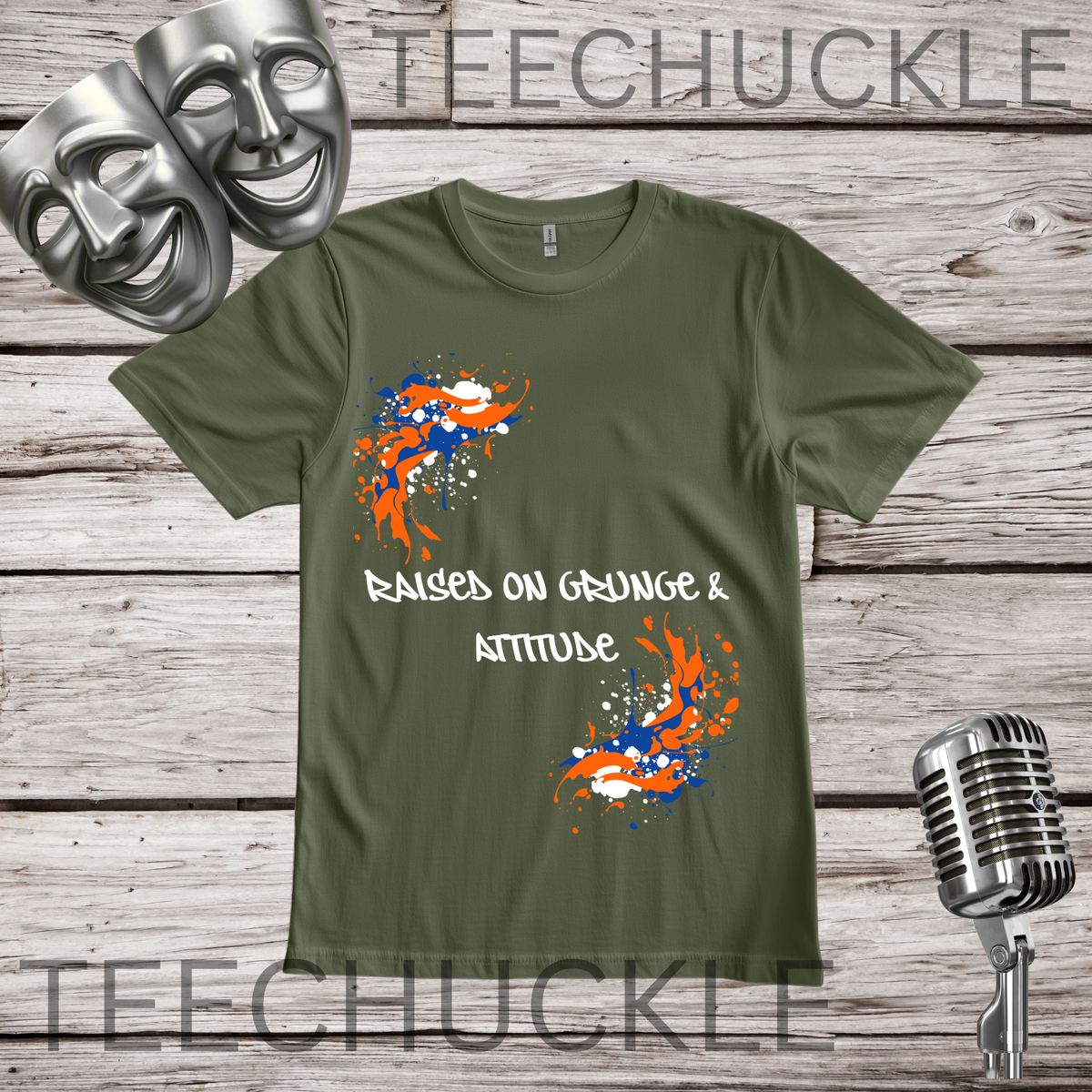 A green graphic t-shirt displayed on a rustic wooden background, styled with two silver comedy masks, a vintage microphone. The t-shirt features orange and blue paint splatters and the white text 'RAISED ON GRUNGE & ATTITUDE'.