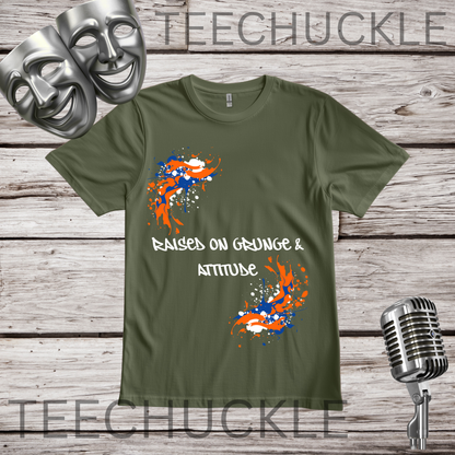 A green graphic t-shirt displayed on a rustic wooden background, styled with two silver comedy masks, a vintage microphone. The t-shirt features orange and blue paint splatters and the white text 'RAISED ON GRUNGE & ATTITUDE'.