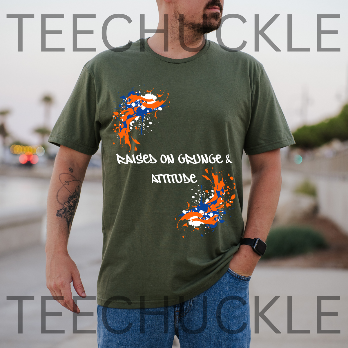 A man wearing a green graphic t-shirt with orange and blue paint splatter designs and the text 'RAISED ON GRUNGE & ATTITUDE', and is set against a blurred outdoor background.